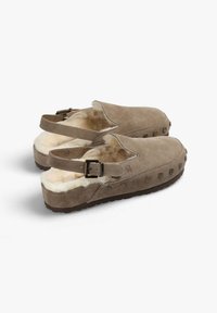 Zuecos de ante marrón con un interior suave, correa ajustable y agujeros redondos a lo largo del lateral. Suela de goma plana para mayor comodidad y estabilidad.