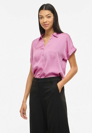 Femeie purtând o bluză roz din satin cu mâneci scurte, băgată într-o pereche de pantaloni negri cu talie înaltă, stând cu o mână în buzunar.