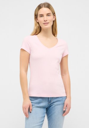 Jeune femme aux longs cheveux blonds portant un t-shirt rose clair à col en V et manches courtes et un jean bleu clair, debout devant un fond uni.