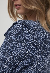 Blouse à motifs marins avec un col à volants, fabriquée dans un tissu léger au design texturé dans des tons de bleu et de blanc.