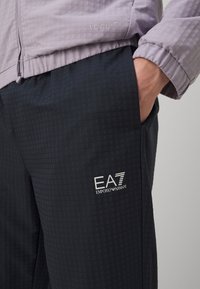 Svijetloplavi zip-up džemper s kariranim uzorkom, u kombinaciji s tamnosivim hlačama koje imaju suptilan uzorak i EA7 logotip.