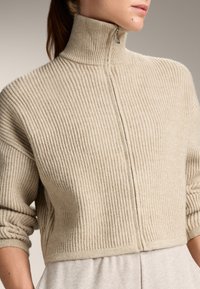Beige Rippstrickpullover mit hohem Kragen und halbem Reißverschluss. Verfügt über ein kurz geschnittenes Design und aufgerollte Ärmel für eine lockere Passform.