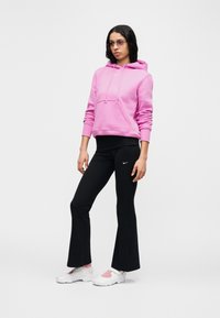 Rosa hettegenser med snorer og lomme, svarte tettsittende bukser med Nike-logo, og hvite sko med rosa detaljer. Modellen står mot en hvit bakgrunn.