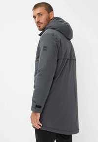 Uomo che indossa un cappotto invernale grigio con cappuccio, mentre guarda oltre la spalla su uno sfondo bianco uniforme.