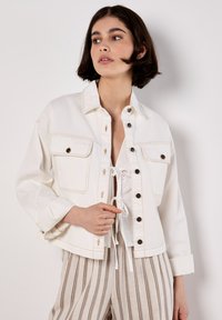 Weiße Jeansjacke mit Brusttaschen, Knopfverschluss und kontrastierenden Nähten. Das Modell trägt ein weißes Oberteil und gestreifte Hosen.