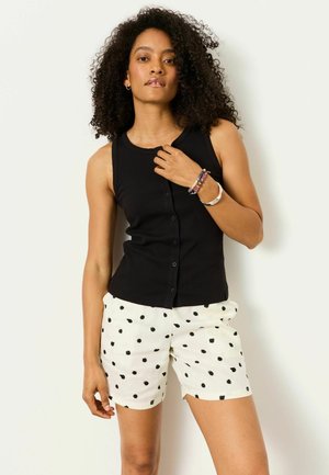 Žena s kučeravými vlasmi, oblečená v čiernom bezrukávovom topu na gombíky, bielych šortkách s čiernymi bodkami a náramkami na zápästí.