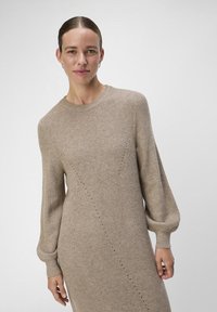 Beige gebreide jurk met lange, puffed mouwen, ronde halslijn en diagonale naaddetails. De textuur is geribbeld en zacht.
