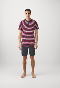 Striped short-sleeve shirt in navy, maroon, and white featuring a button placket, paired with dark shorts. Model stands barefoot against a plain background.