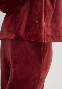 Samt-Loungewear-Set in tiefrot, bestehend aus einem lässigen Oberteil und Hose mit Seitentasche, glatter Textur und subtiler Logodetails.