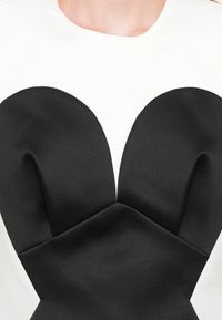 Corset din satin negru cu cupe proeminente în formă de inimă, țesătură albă în partea de jos. Textură netedă și detalii unghiulare la cusături vizibile.