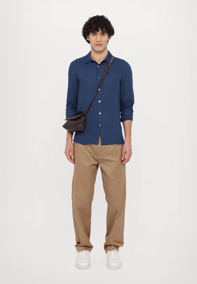Marineblauw overhemd met lange mouwen, tan kleurige wijde broek, witte sneakers en een bruine schoudertas. Glad materiaal, relaxte pasvorm.