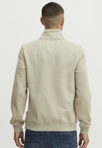 Sudadera beige con cuello alto, puños y dobladillo acanalados; textura suave; combinada con jeans azules; sin patrones o gráficos visibles.