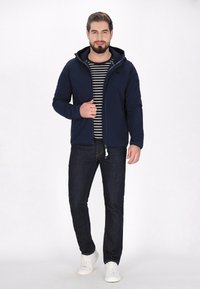 Mornarska zip-srajca s kapuco, kombinirana z črtasto majico, temnimi kavbojkami in belimi športnimi copati. Gladek material, sproščen dizajn, funkcionalni žepi.