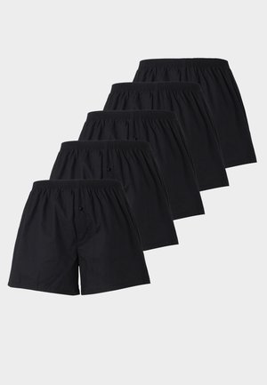 Bărbat musculos, fără tricou, stând desculț, purtând chiloți boxer negri pe un fundal simplu și deschis.
