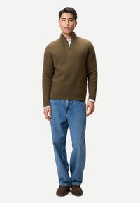 Junger Mann trägt einen olivgrünen Pullover mit halbhohem Reißverschluss, hellblaue Jeans und braune Schuhe, steht vor einem schlichten weißen Hintergrund.