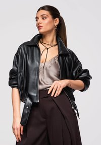 Jonge vrouw met een zwarte leren jas, taupe kanten hemdje, donkere broek en een dunne zwarte ketting, poserend tegen een effen achtergrond.