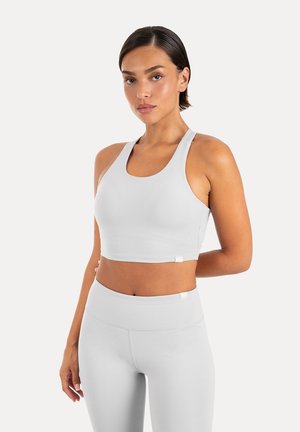 Frau trägt ein hellgraues ärmelloses Crop-Top und passende hochtaillierte Leggings und steht vor einem schlichten Hintergrund.