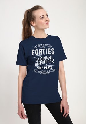 Ung kvinde med brunt hestehale iført mørkeblå t-shirt med hvid tekst: "Bygget i fyrrerne, original og urestaureret, nogle dele fungerer stadig."