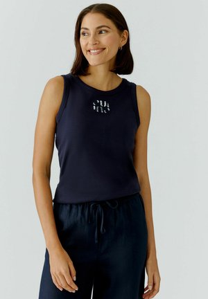 Vrouw met donker haar tot op de schouders, gekleed in een donkerblauw mouwloos topje met kleine witte tekst en donkerblauwe broek met trekkoord, glimlachend en naar rechts kijkend.