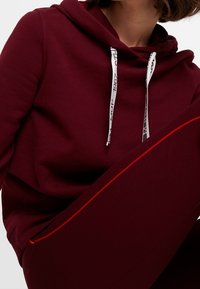 Burgundská mikina s bílými stahovacími šňůrkami a černým textem; měkká, strukturovaná tkanina; spárovaná s odpovídajícími burgundskými kalhotami s červeným akcentem.