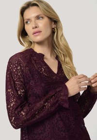 Blusa in pizzo bordeaux con scollatura a V, colletto a rouches e lunghi manicotti a campana con motivi intricati e sezioni trasparenti.