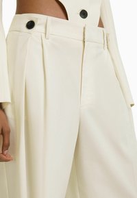 Cream-colored tailored trousers with pleats, featuring black buttons and a smooth, soft texture. Front pockets are visible.