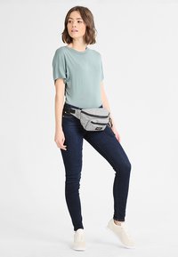 Šedá látková ledvinka se dvěma zipovými kapsami, nošená kolem pasu. Modelka má na sobě světle zelenou košili a tmavé úzké džíny, stojí na neutrálním pozadí.