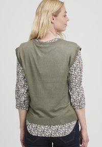 Oliven grøn ærmeløs top med ribbet kant, lagt over en bluse med blomsterprint, der har trekvartærmer og en buet kant.