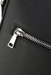 Superficie in pelle nera con texture, con una cerniera in metallo argentato parzialmente chiusa e un tirante rettangolare su una borsa.