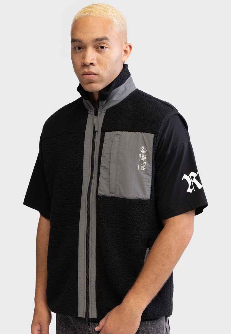 Gilet en polaire noire avec des accents gris, fermeture éclair sur toute la longueur à l'avant et une poche poitrine. T-shirt noir à manches courtes en dessous. Texte imprimé sur la poche.