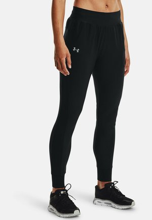 Osoba nosząca czarne dopasowane sportowe spodnie dresowe z logo Under Armour oraz czarne buty do biegania z białymi podeszwami na jednolitym tle.