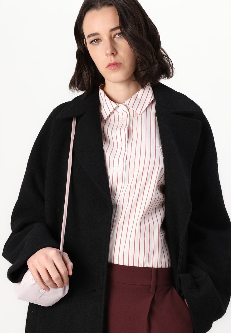 Schwarzer Wollmantel mit weiten Ärmeln, über einem weißen Hemd mit roten Streifen. Das Modell hält eine kleine rosa Lederhandtasche. Burgunderfarbene Hosen runden das Outfit ab.