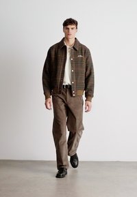 Jachetă bomber cu carouri maro, cu manșete ribbate, purtată cu un polo de culoare deschisă și pantaloni maro, asortată cu pantofi negri pe un fundal neutru.