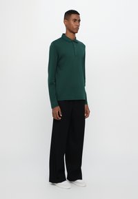 Grønn langermet polo skjorte med krage og tre knapper, kombinert med løstsittende svarte bukser og hvite sneakers. Enkel, ren design.