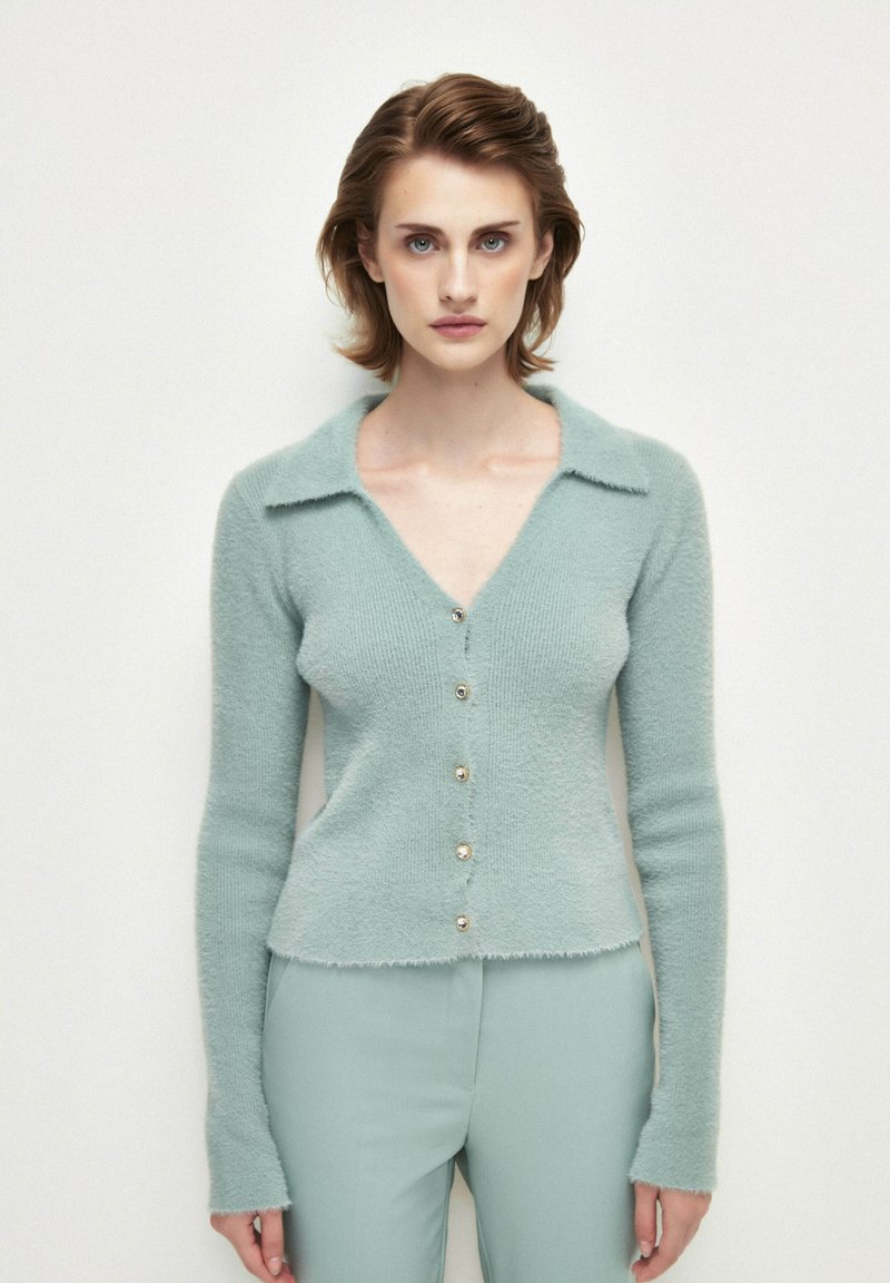 Ljus teal, långärmad cardigan med v-ringning, texturerat material och fem gyllene knappar framtill. Fransade kanter på nederkanten och kragen.