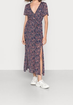 Frau trägt ein marineblaues, geblümtes Hemdblusenkleid mit tiefem V-Ausschnitt, Seitenschlitz, kurzen Ärmeln und weißen klobigen Turnschuhen.