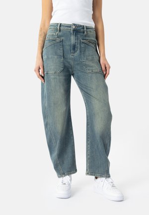 Osoba ubrana w luźne niebieskie dżinsy z dużymi przednimi kieszeniami i białe tenisówki, stojąca na jednolitym tle.