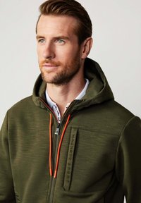 Giacca verde con cappuccio in un tessuto texturizzato, dotata di cordini arancioni e una tasca sul petto con zip, con il logo "HACKETT SPORT" impresso.