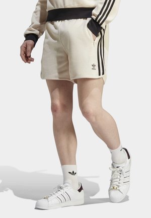 Persoon draagt beige Adidas short en bijpassend sweatshirt met zwarte strepen, witte Adidas sokken en witte sneakers met zwarte strepen.