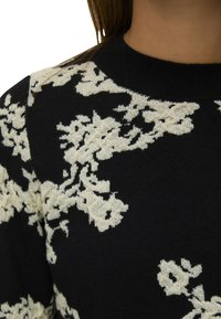 Pull en maille noire avec des motifs floraux crème, col arrondi et tissu texturé. Vue rapprochée de la partie supérieure.