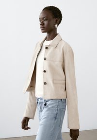 Beige mocka-jacka med krage, framsida med knappar och sidofickor, bärs över en vit topp och ljusblå jeans.