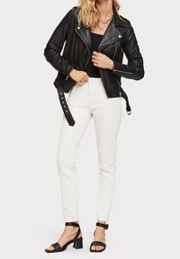 Czarna skórzana kurtka motocyklowa z srebrnymi zamkami, w połączeniu z białymi dopasowanymi dżinsami i czarnymi sandałami na słupku z plecionymi paskami.
