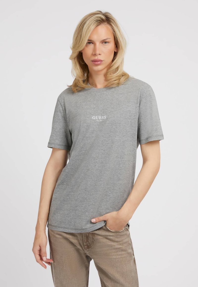 Grå t-shirt med korte ærmer og rund halsudskæring, prydet med et lille hvidt "GUESS" logo foran, sat sammen med beige bukser.