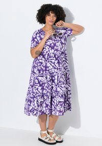 Langes lila Tie-Dye-Maxikleid mit kurzen Ärmeln und gestuften Rüschen. Es hat einen runden Ausschnitt und einen leichten, strukturierten Stoff. Weiße Sandalen.