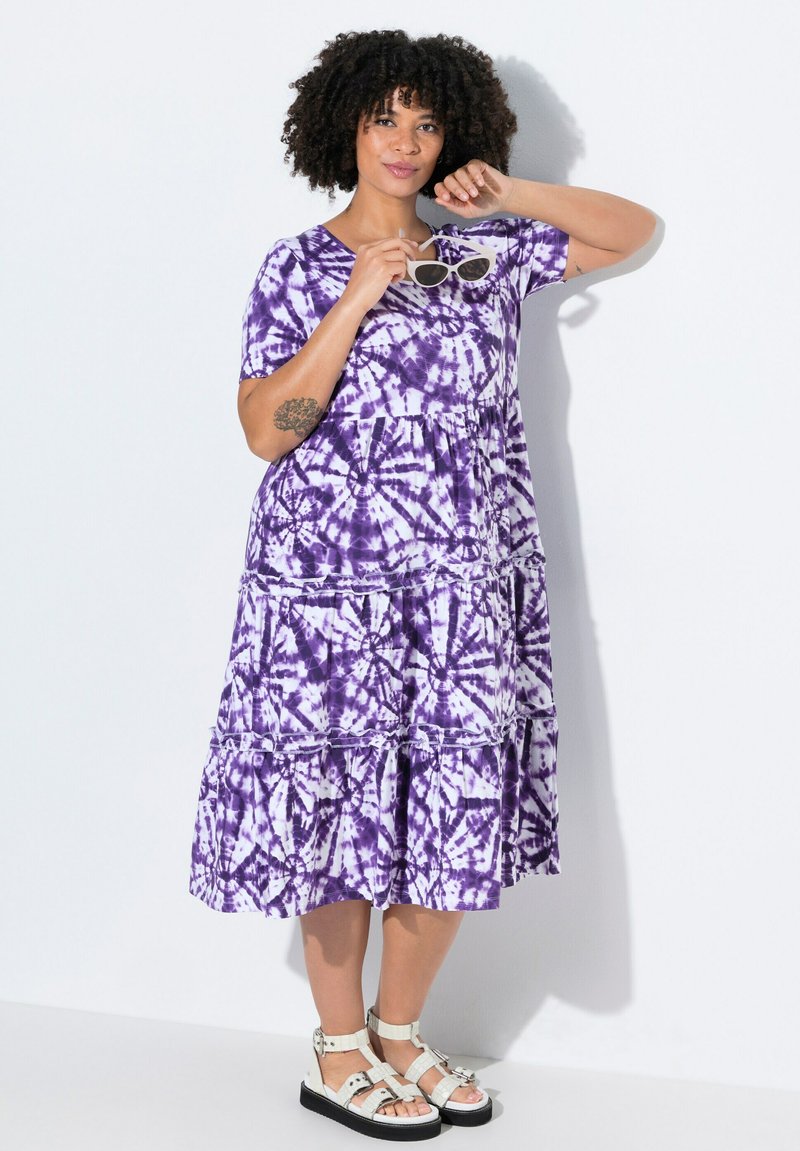 Langes lila Tie-Dye-Maxikleid mit kurzen Ärmeln und gestuften Rüschen. Es hat einen runden Ausschnitt und einen leichten, strukturierten Stoff. Weiße Sandalen.