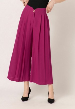 Kvinde iført magenta bukser med høj talje og vide ben med læg, kombineret med sorte højhælede sko og en sort top, hænder på hofterne.