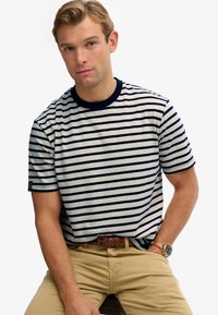 Stribet t-shirt i off-white og marineblå, kortærmet, med en ribbet rund hals. Sættes sammen med tanfarvede bukser og et flettet brunt bælte.