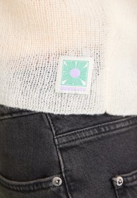Maglione di colore crema lavorato a maglia con una trama larga, caratterizzato da una toppa logo verde e viola. Jeans grigio scuro con bottoni in metallo.