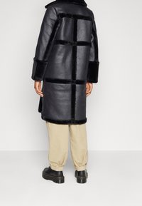 Abrigo de cuero negro con acentos de piel sintética, que presenta un patrón de cuadrícula. La modelo lleva pantalones crema y zapatos negros de suela gruesa. Primer plano de los materiales visible.