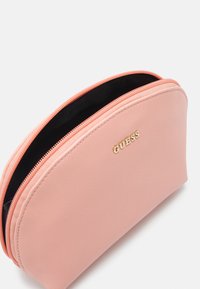 Pochette cosmetica sintetica color pesca con forma arrotondata, chiusura con doppia zip e logo dorato "GUESS". Fodera interna nera visibile.